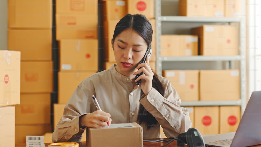 Asian woman working at online store warehouse using mobile phone talking with customer, online e-commerce retail small business concept