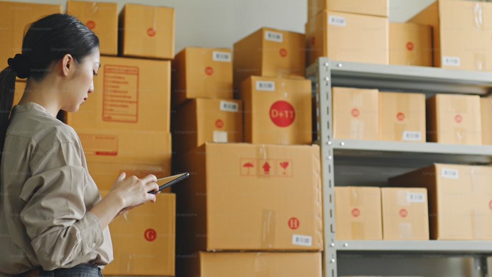 Asian woman working at online store warehouse checking inventory stock parcel boxes on shelves, online e-commerce retail small business concept