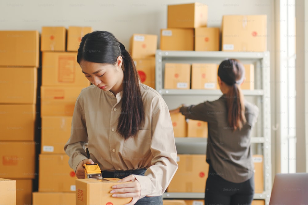Asian woman working at online store warehouse packing product to parcel cardboard boxes, online e-commerce retail small business concept