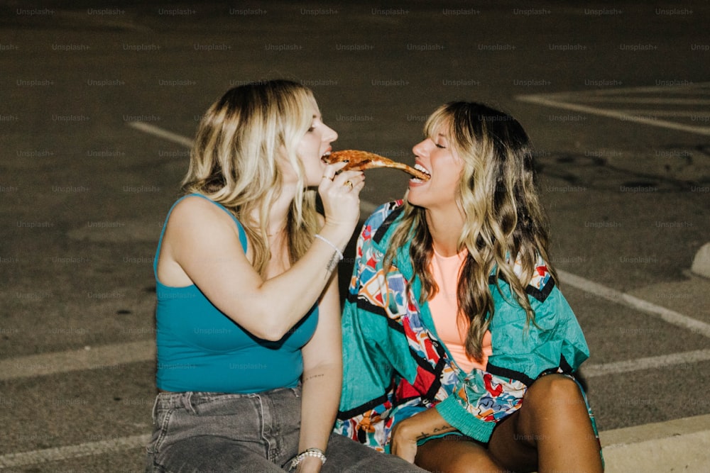 two women sitting on the ground eating pizza