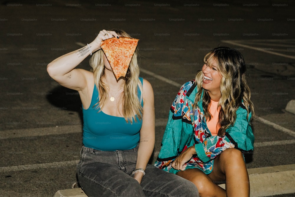 two women sitting in a parking lot, one with a slice of pizza on her