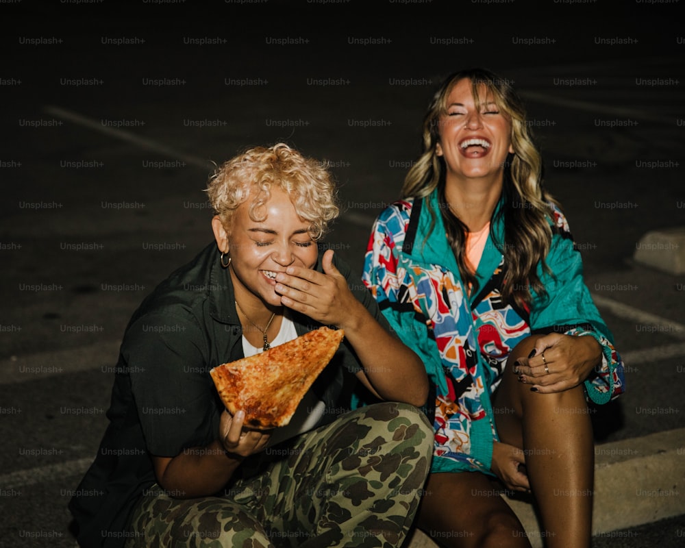 a couple of women sitting next to each other eating a slice of pizza
