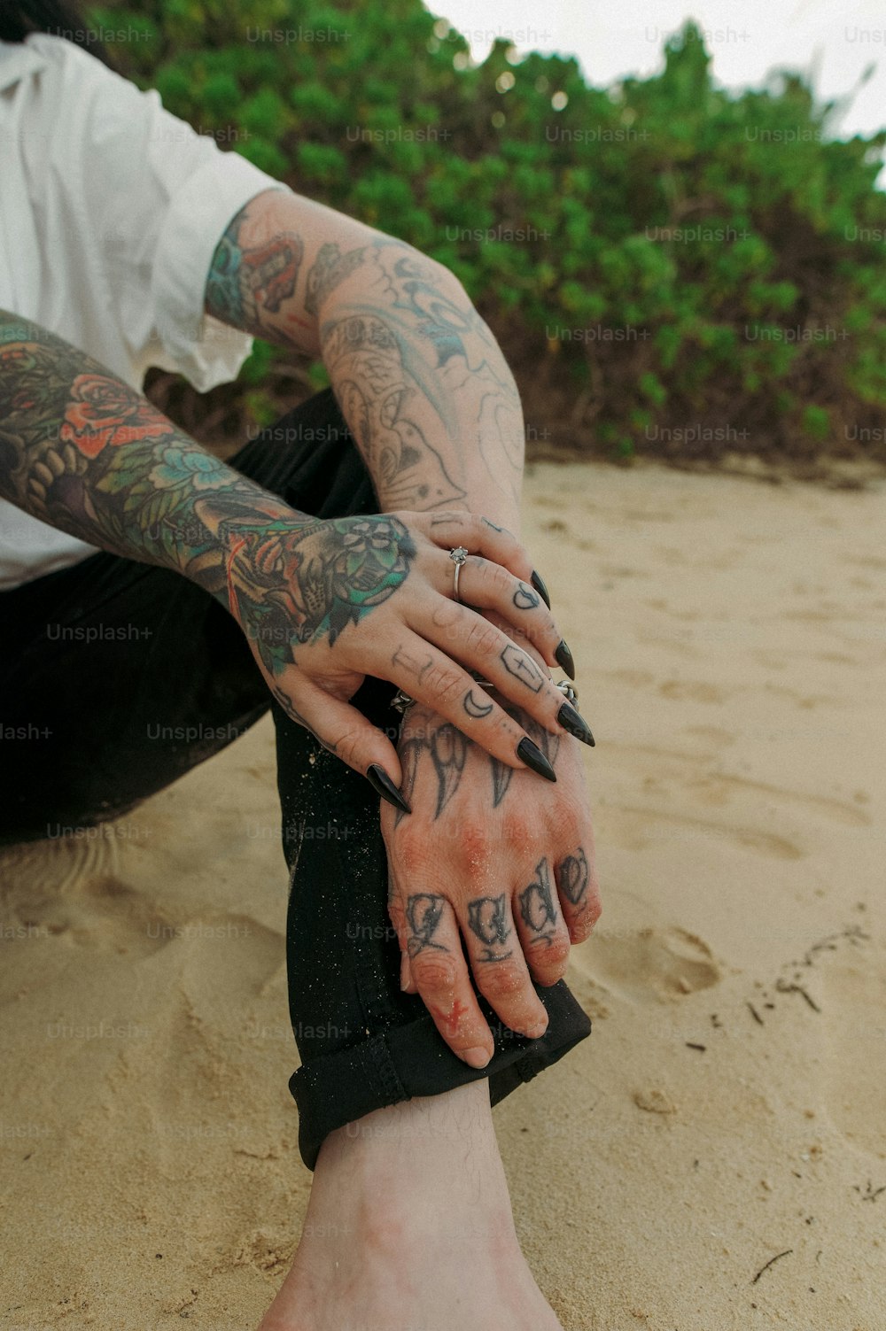 a tattooed man sitting on the beach with his hands on his knees