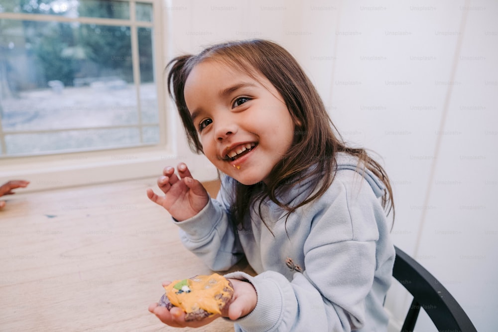 Ein kleines Mädchen, das mit einem Stück Essen in der Hand an einem Tisch sitzt
