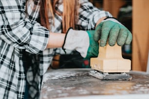 a woman in a plaid shirt and green gloves sanding a piece of wood