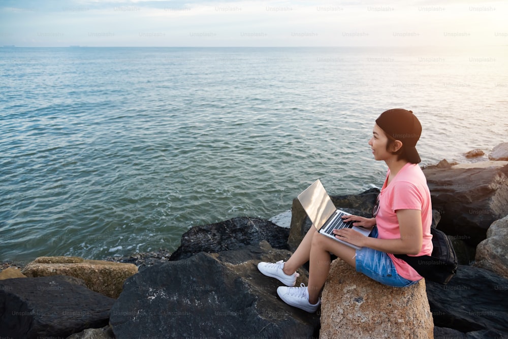 Joven mujer asiática independiente con camisa rosa sentada en la roca y trabajando con computadora portátil al aire libre. Fondo del mar y de la puesta del sol.