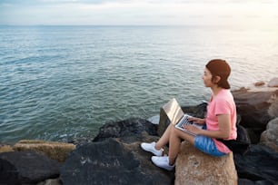 Young Asian Woman Freelance in pink shirt sitting on the rock and working with laptop at outdoor. Sea and sunset background.
