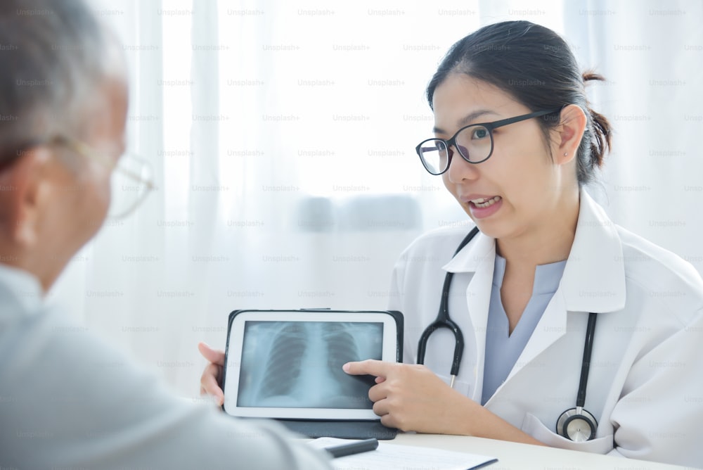 眼鏡をかけている若いアジアの女性医師は、診療所のデジタルタブレットコンピュータで胸部X線写真について年配の男性患者と話し合っています。レントゲン。