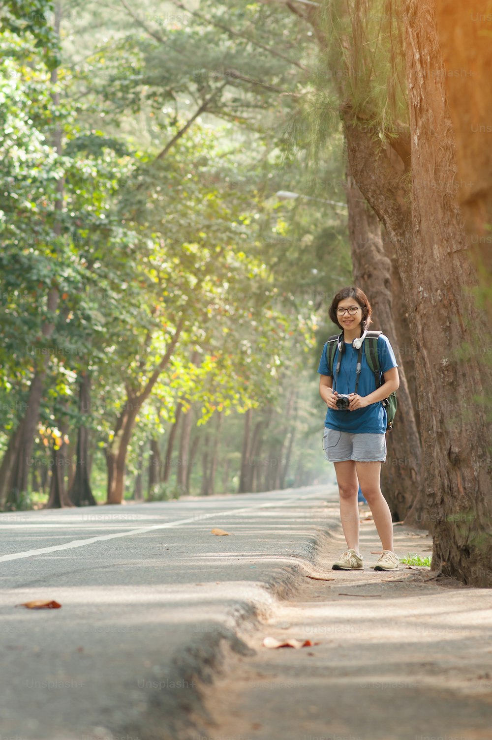 Asian woman hitchhiker with backpack and camera on highway road with sunlight on nature background.