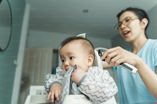 Joven madre asiática cortando el pelo a su pequeño hijo bebé por sí misma en casa, madre china cortando el pelo del niño con cortapelos, peluquero