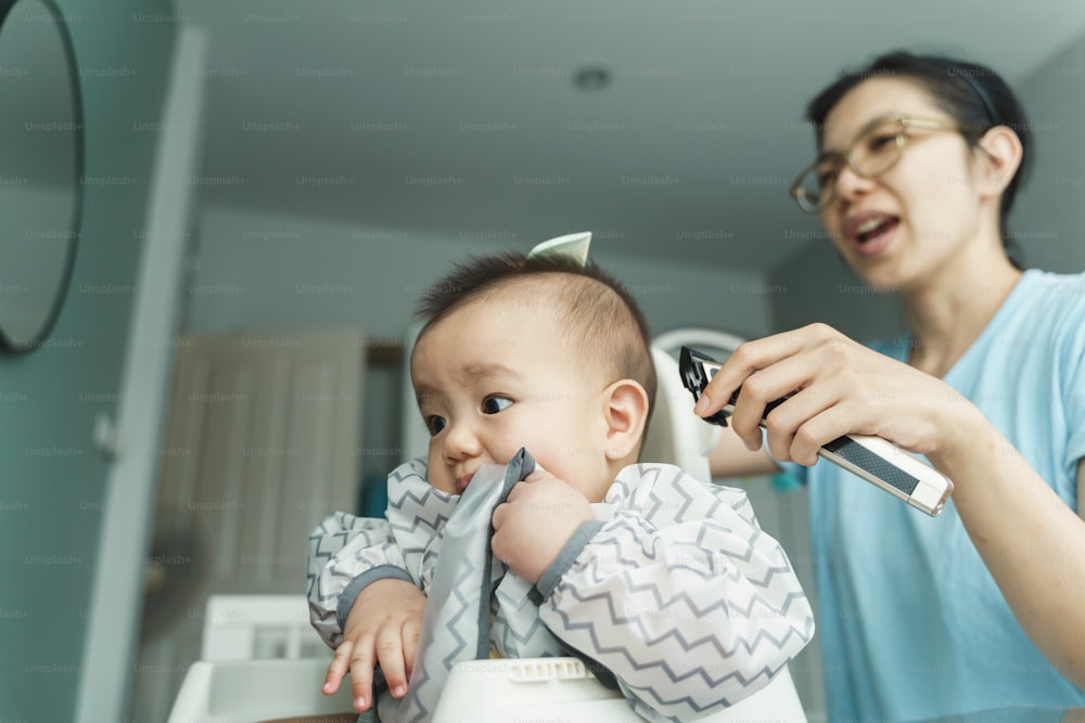 Asian Young Mother cutting hair to her little baby son by herself at home, Chinese Mom cutting hair of child with hair clipper, Hairdresser