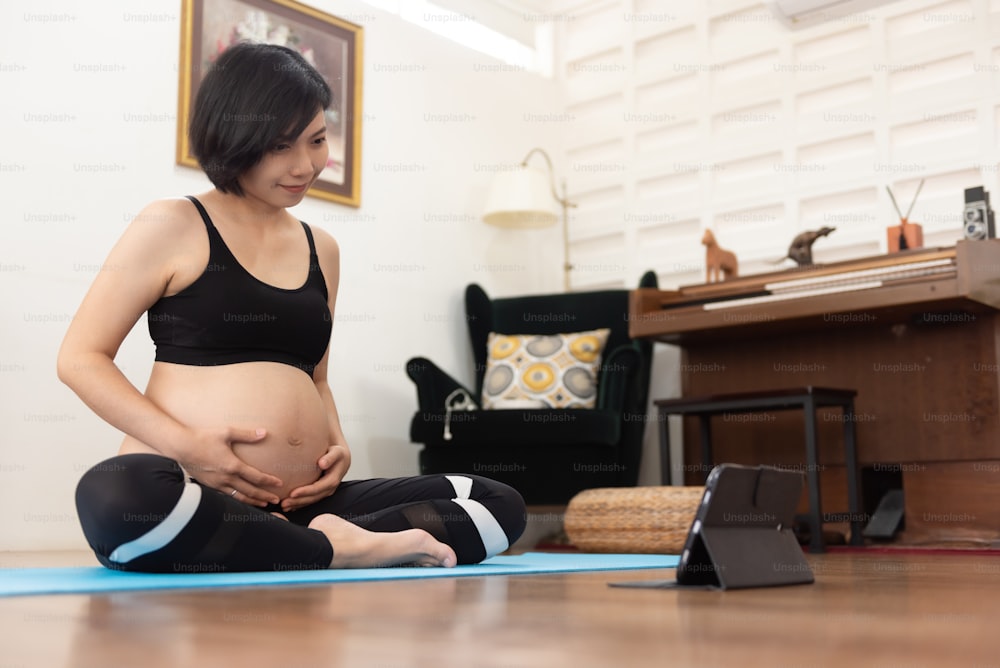 Femme enceinte asiatique pratiquant le yoga en ligne avec une tablette numérique à la maison.