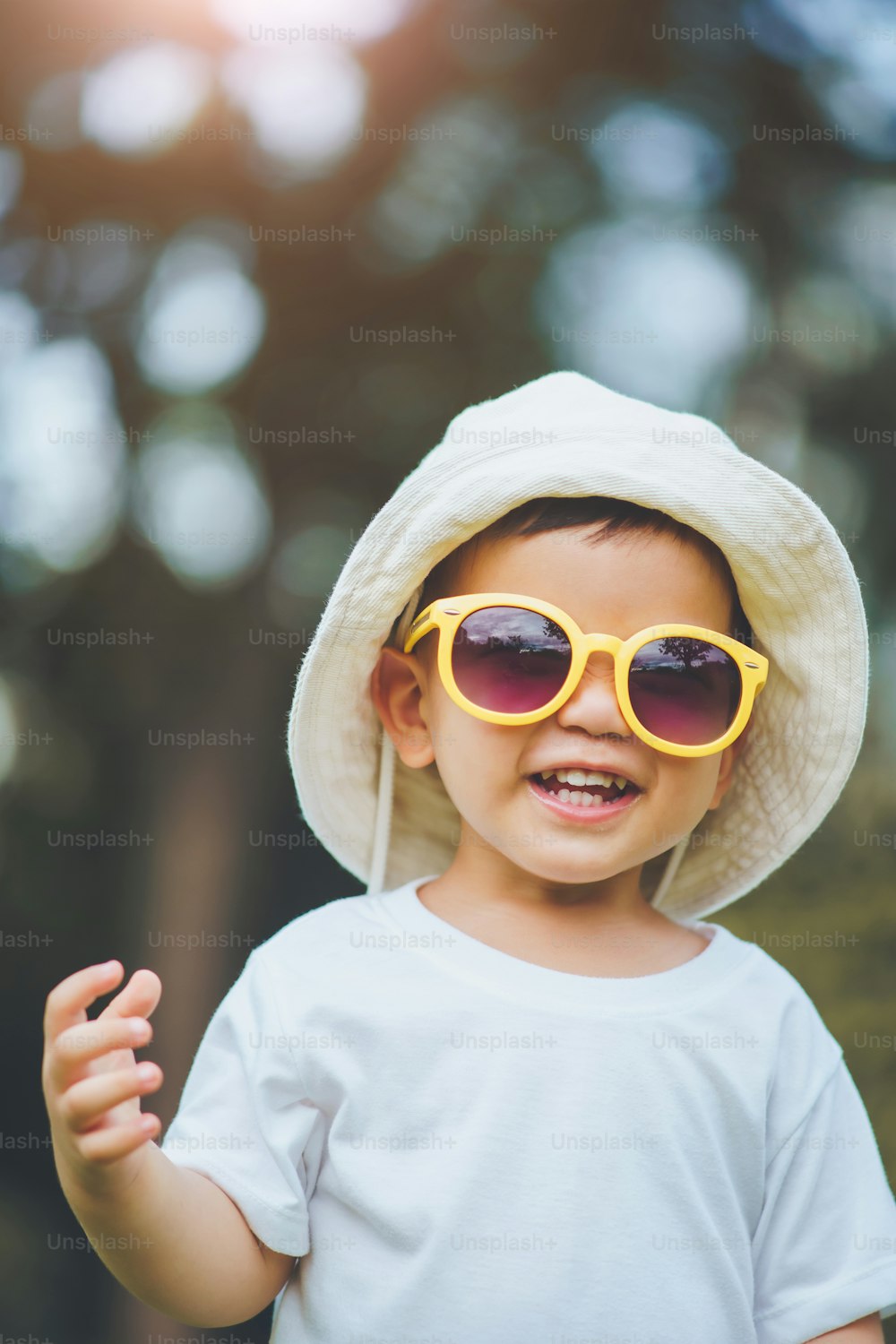 Ritratto di ragazzino asiatico felice che indossa cappello e occhiali da sole gialli che guardano la macchina fotografica all'aperto
