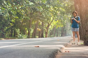 Asian woman hitchhiker with backpack looking camera on highway road with sunlight on nature background.