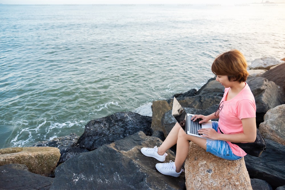 Joven mujer asiática independiente con camisa rosa sentada en la roca y trabajando con computadora portátil al aire libre. Fondo del mar y de la puesta del sol.