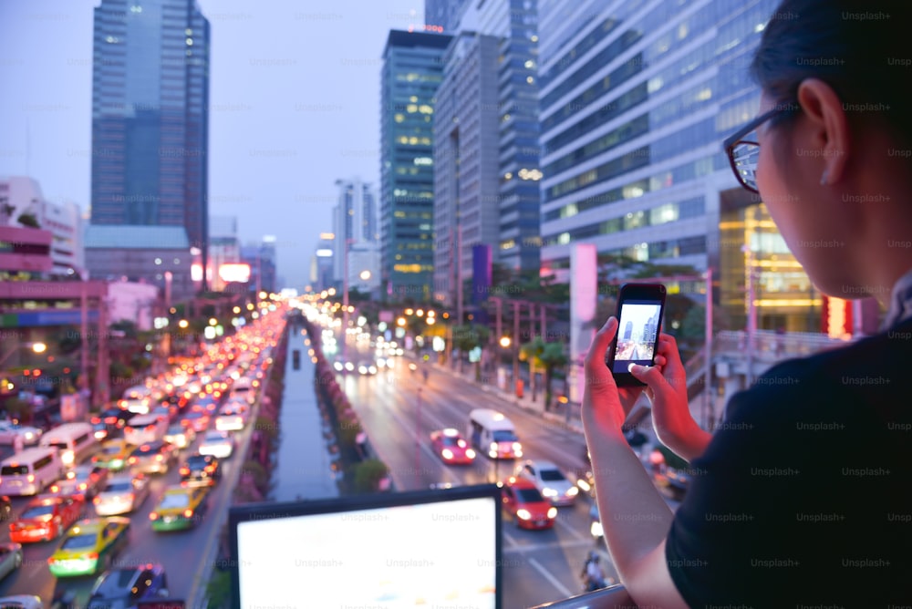 Mulher asiática tirando foto com seu smartphone no engarrafamento e edifícios com fundo de semáforo noturno na cidade. Espaço de cópia.