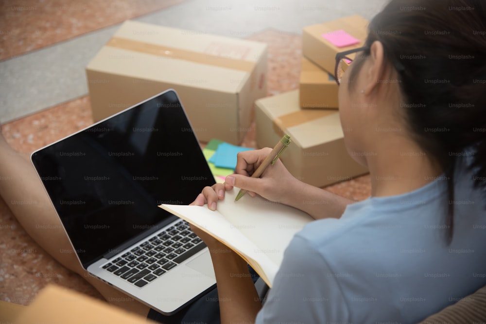 Young Asian woman looking to order details and checking supplies with notebook, laptop and mobile phone in her small warehouse. Own business