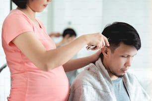 Young Pregnant Woman cutting man hair with clipper during pandemic virus at home. Asian husband getting haircut from his wife during quarantine. Lifestyle of Couple in family barbershop