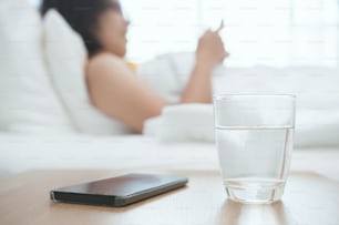 �スマートフォンを前景にした女性のグラスの水を接写します。美しいアジアの女の子は、彼女のスマートフォンを見て、ベッドに横たわっています。