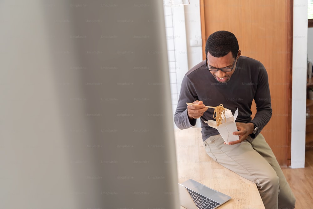 Uomo afroamericano che fa videochiamata con i suoi amici sul laptop mentre mangia una tazza di noodle istantaneo con bacchette. Maschio nero sorridente che ha soba dalla scatola da asporto mentre la quarantena covid-19 a casa