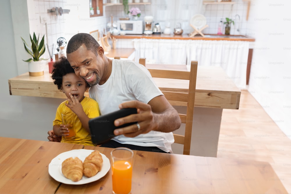 Happy African Father and son taking selfie photo with smartphone during lunch time in dining and kitchen room. Portrait smiling Dad and little kid boy making video call with mobile phone together in morning at home. Black Family having fun