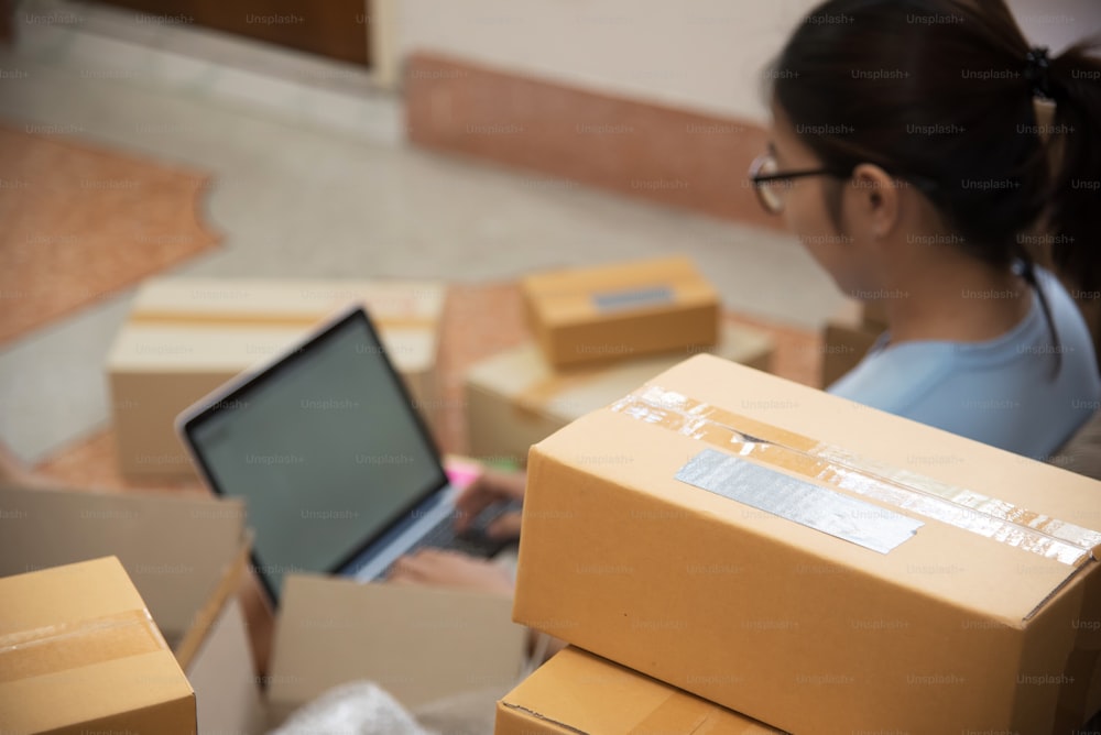 Selective focus on box on foreground. Young Asian woman using a laptop in her small warehouse. Own business