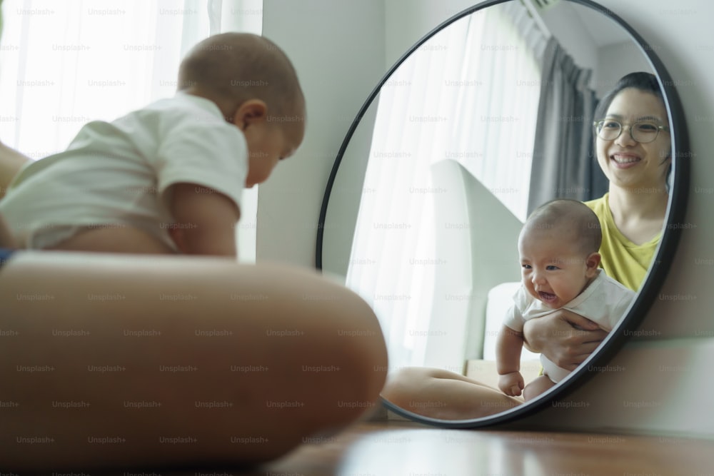 Playful Asian mother and newborn baby boy having fun sitting looking at the mirror at home. Cheerful Young Mom smiling embracing playing with her son on the floor together.