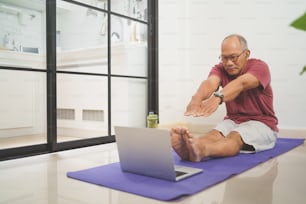 Homem idoso asiático ativo olhando sala de aula on-line no laptop enquanto faz exercícios de alongamento em casa