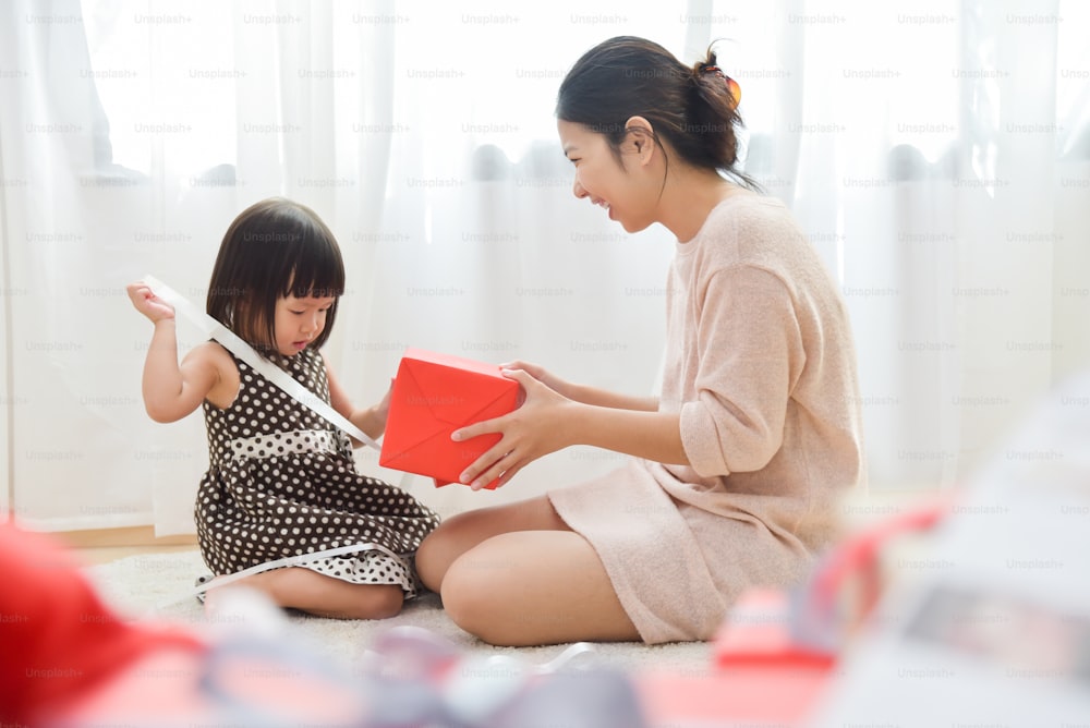 Asian Young Mother is Looking to Little girl packt eine rote Geschenkbox in einem weißen Raum für Geburtstag, Weihnachten und Neujahr aus. Speicherplatz kopieren. Fröhliche Familienfeier.