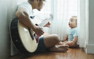 Asian Father sitting singing and playing acoustic guitar with little Cute baby infant boy together on wooden floor, Focus on child