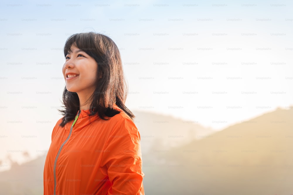 Porträt asiatische Frau in orangefarbener Jacke lächelt und steht über Berghintergrund. Vision, erfolgreich, positive Emotion.