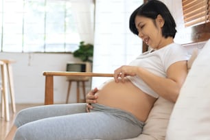 중국 또는 일본 여성 소파에 앉아 임신한 배를 연주, 젊은 여자 기대 아기 그녀의 배를 만지고