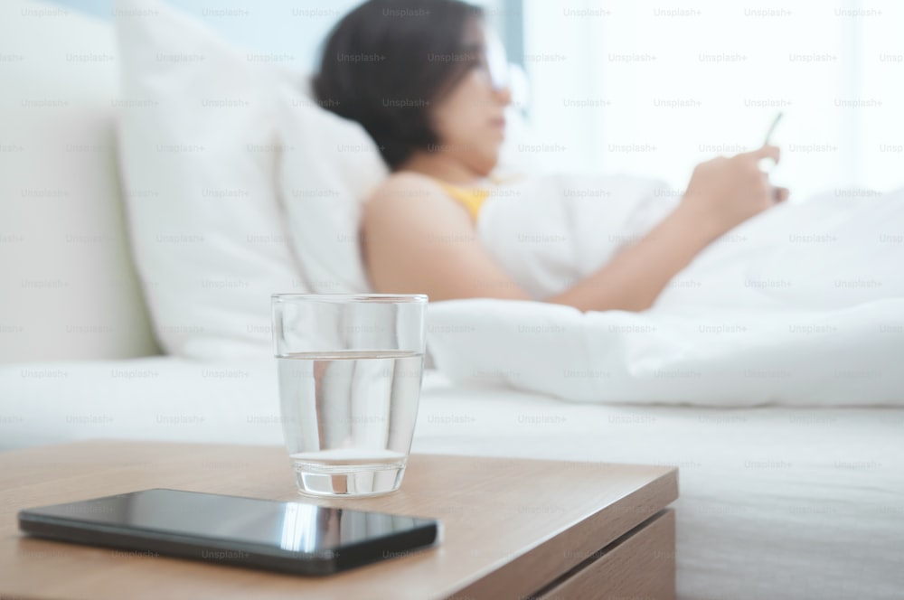 Gros plan sur le verre d’eau d’une femme avec un smartphone au premier plan. Belle fille asiatique regardant son téléphone intelligent et allongée sur le lit.
