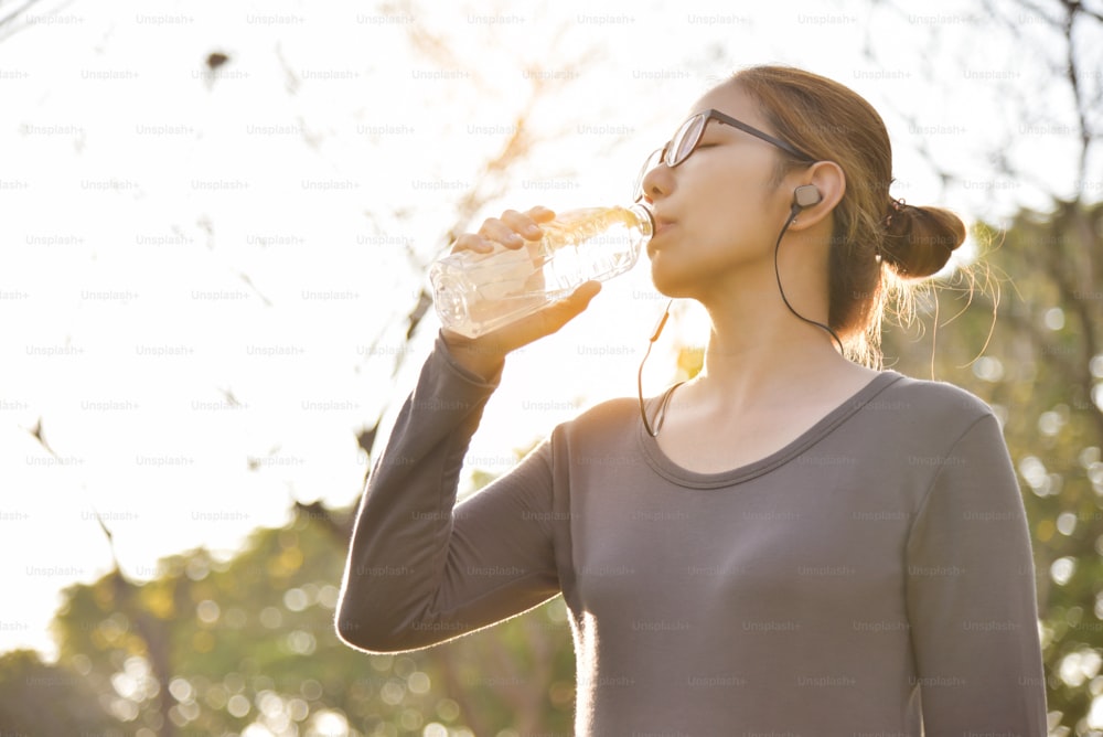 회색 운동복을 입은 젊은 아시아 여성이 공원에서 운동하는 동안 휴식을 취하고 물을 마시고 있다. 아침에 야외에서 이어폰으로 음악을 듣는 여자.  건강 관리 개념입니다.  일몰과 햇빛. 공간을 복사합니다.