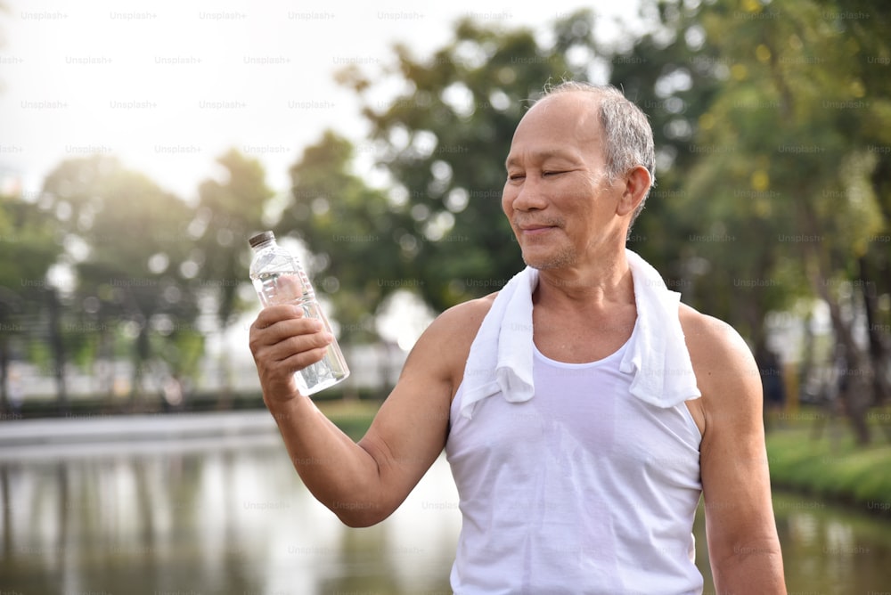 Asiatischer älterer Mann, der eine Flasche Wasser zum Trinken hält, während er im Park im Freien trainiert.