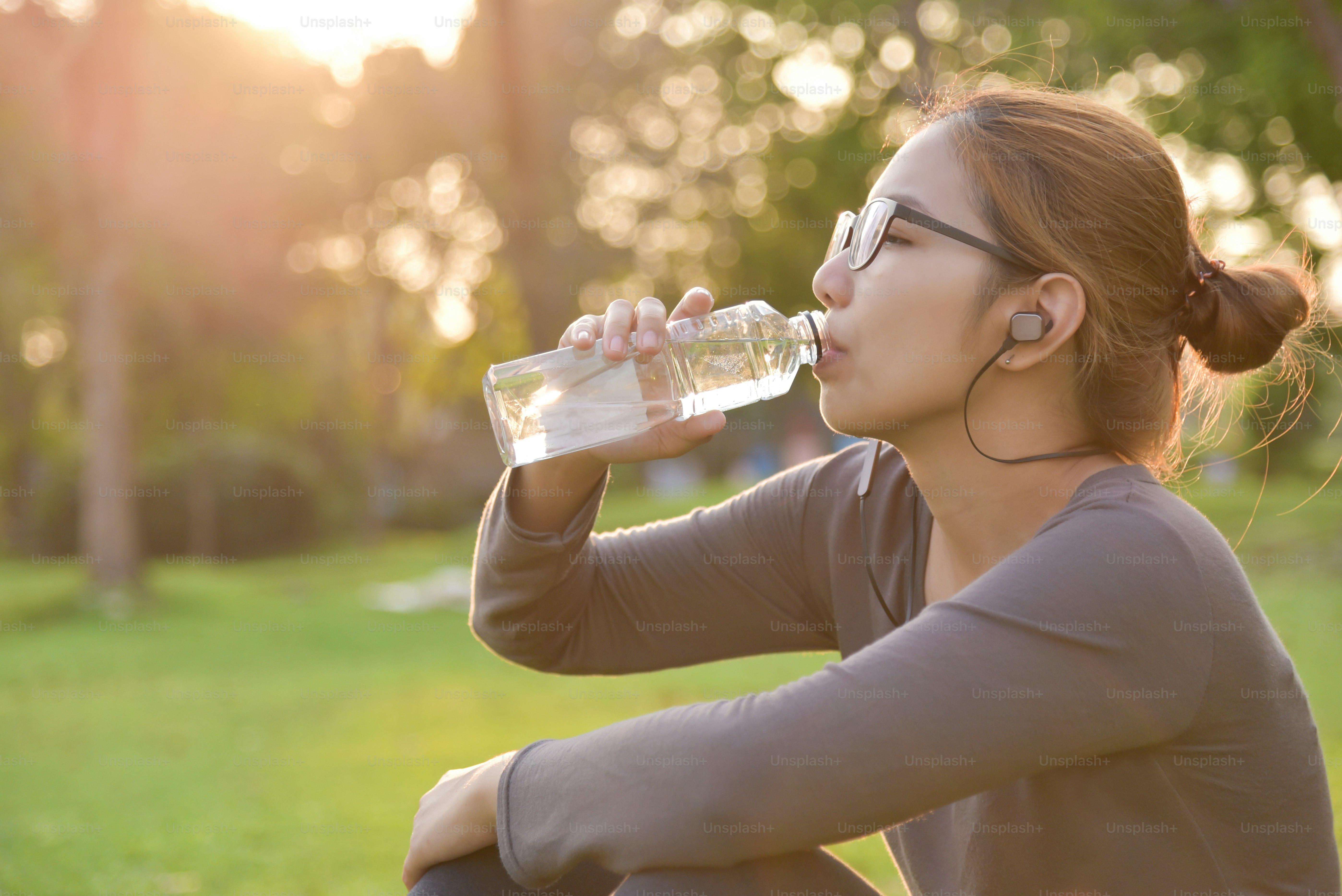 Young Asian female  in Grey sportswear is resting and drinking water while exercise at park. Woman listening to the music with earphones at outdoors on morning.  Health care concept.  Sunset and Sunlight. Copy space.