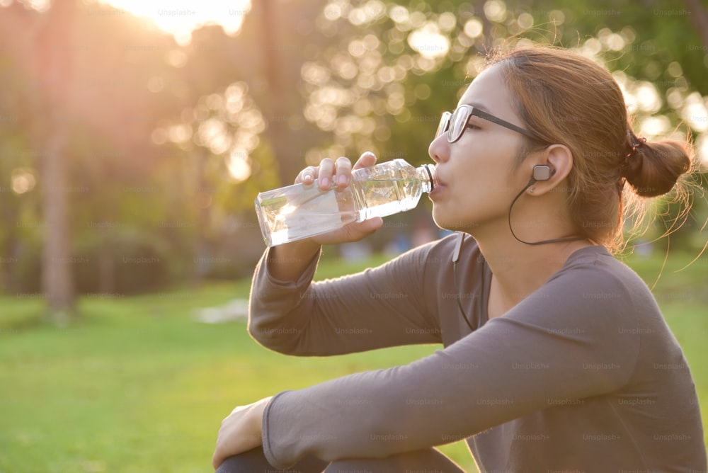 Une jeune femme asiatique en vêtements de sport gris se repose et boit de l’eau tout en faisant de l’exercice au parc. Femme écoutant de la musique avec des écouteurs à l’extérieur le matin.  Concept de soins de santé.  Coucher de soleil et lumière du soleil. Espace de copie.