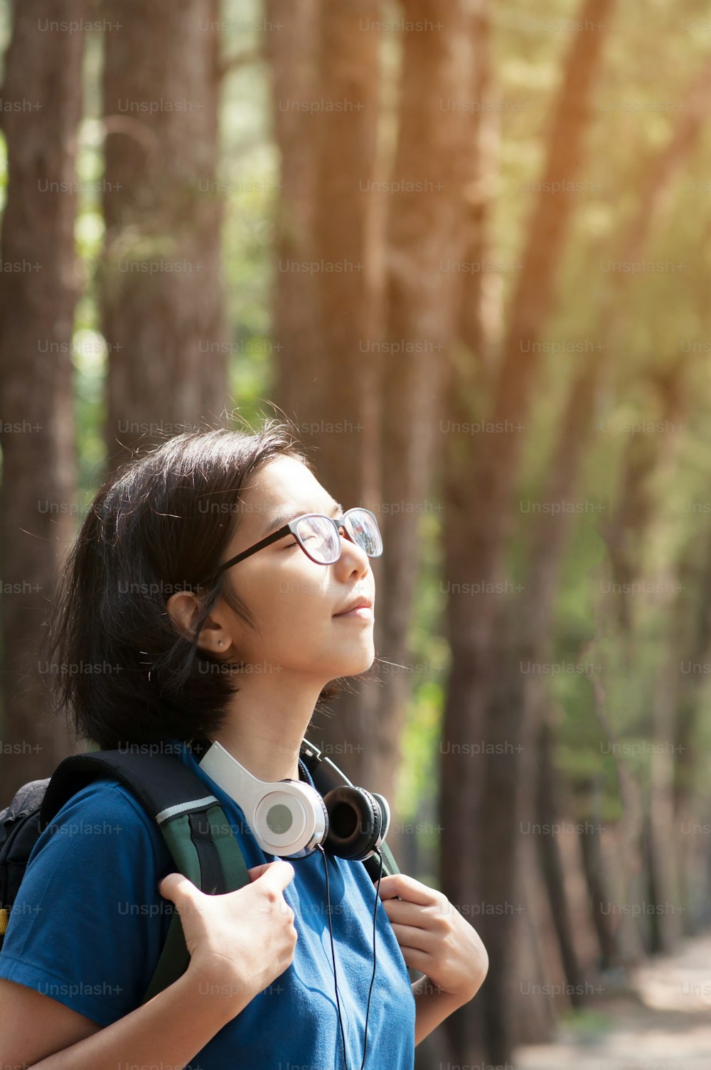 Óculos asiáticos mulher caminhante com mochila respirando natureza ar fresco. Menina bonita com fone de ouvido sorrindo no fundo da natureza.