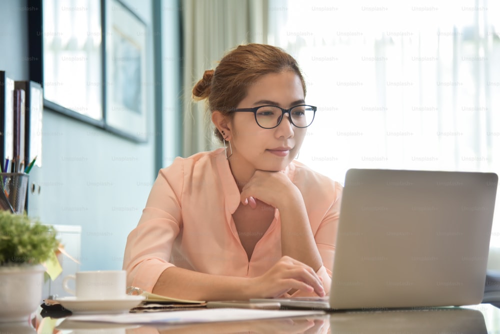 幸せな若いアジアの女性クリエイティブデザイナーは、職場でラップトップコンピュータで作業する眼鏡をかけています。自宅でネットショッピングをしている女性。
