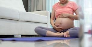 Femme enceinte asiatique assise sur un tapis de yoga touchant son ventre tout en faisant des exercices de yoga.