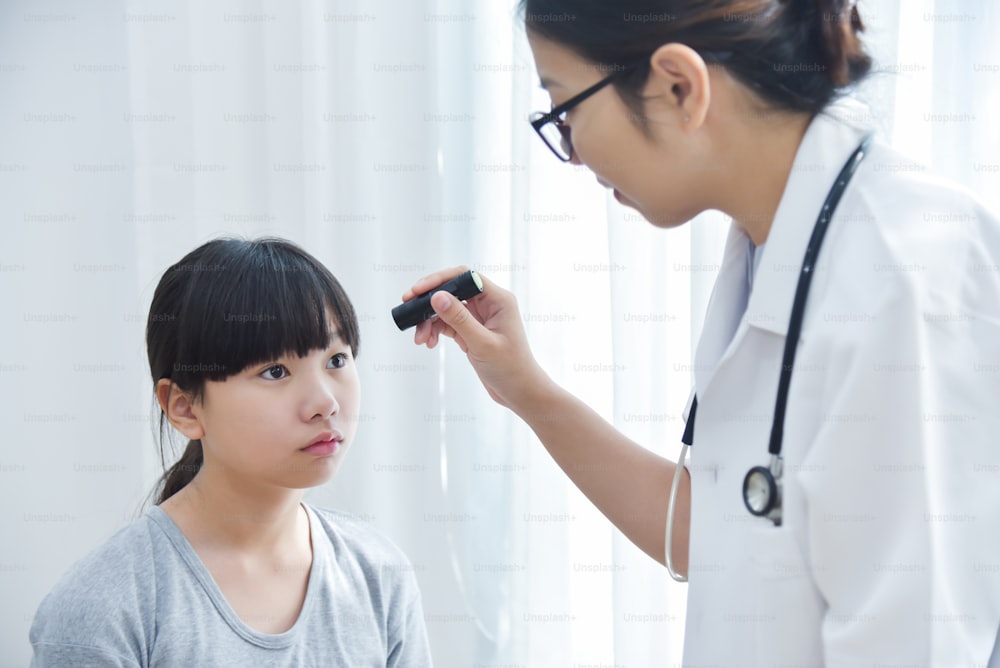 Junge asiatische Ärztin trägt eine Brille, die die Augen eines kleinen Mädchens mit Taschenlampe in der Arztpraxis überprüft.