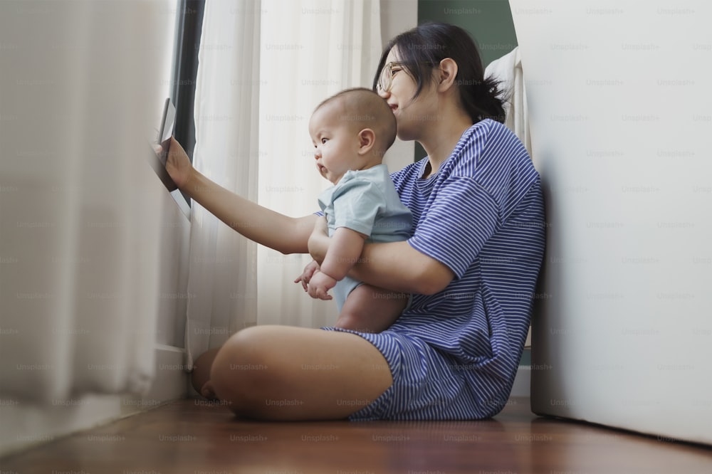 Asian Junge Mutter und neugeborener Junge sitzen auf dem Boden und führen Videoanrufe mit der Familie auf dem Smartphone zu Hause. Happy Smiling Mama und Sohn machen Selfie zusammen mit Handy.