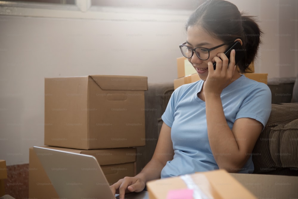 Young Asian woman looking to order details and checking supplies on laptop and mobile phone in her small warehouse. Own business
