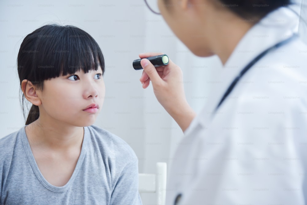 Junge asiatische Ärztin trägt eine Brille, die die Augen eines kleinen Mädchens mit Taschenlampe in der Arztpraxis überprüft.