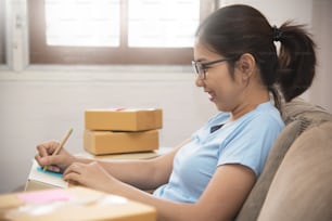 Giovane donna asiatica che controlla le forniture e scrive sulla scatola di cartone nel suo piccolo magazzino. Attività in proprio