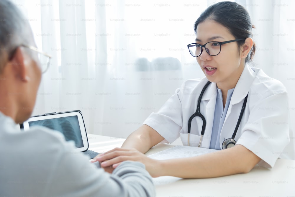 Joven médico asiático tranquilizando al paciente masculino mayor con la mano en el consultorio médico.