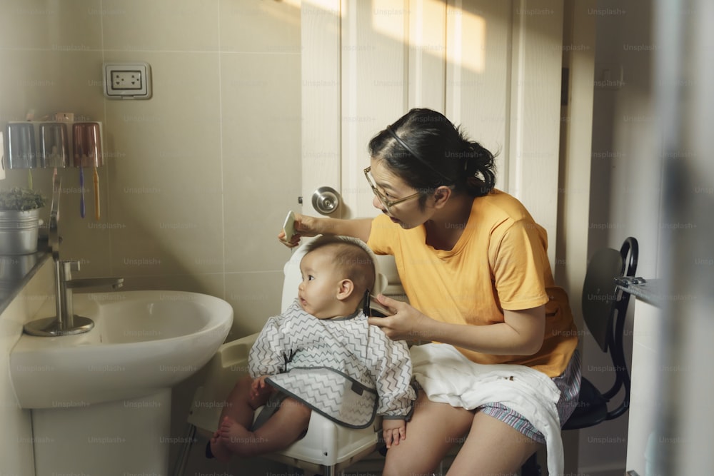 Madre asiática cortando el pelo a su pequeño hijo sola con una cortadora de pelo en el baño de casa.