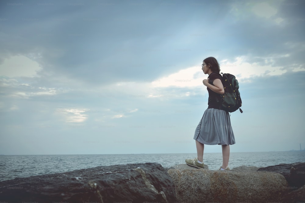 Young Asian woman traveller standing on cliff edge and looking to sea with cloudy sky.