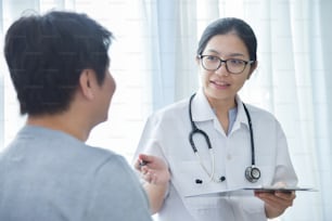 Asian Female Doctor examining and taking note on checklist paper with male patients in medical room.