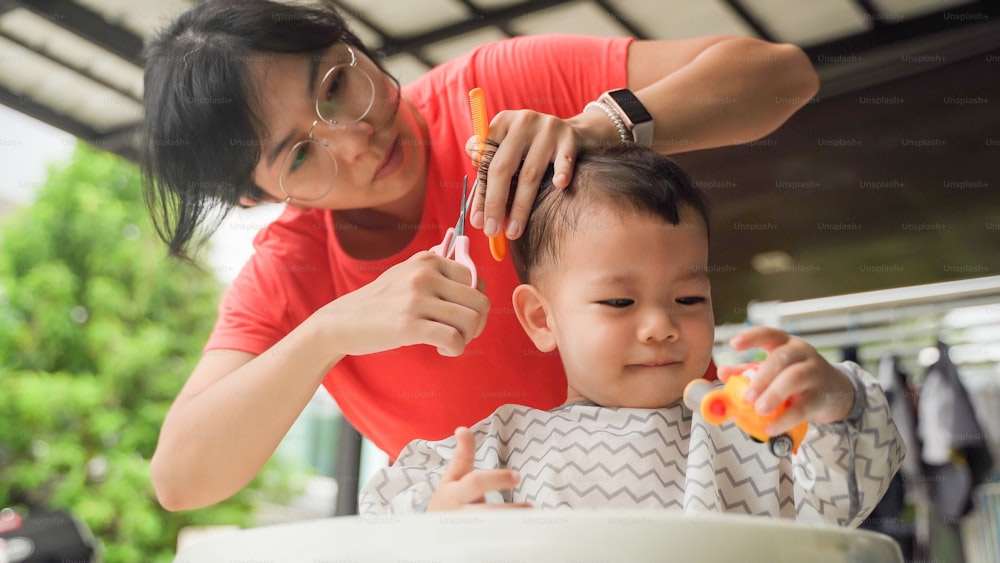 Asiatische Mutter, die ihrem kleinen Sohn zu Hause draußen die Haare schneidet, entzückender Junge, der Spaß hat.